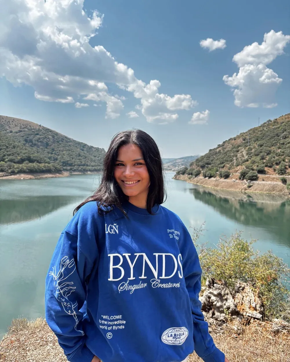 Woman by a lake wearing a blue BYNDS Singular Creatures sweatshirt