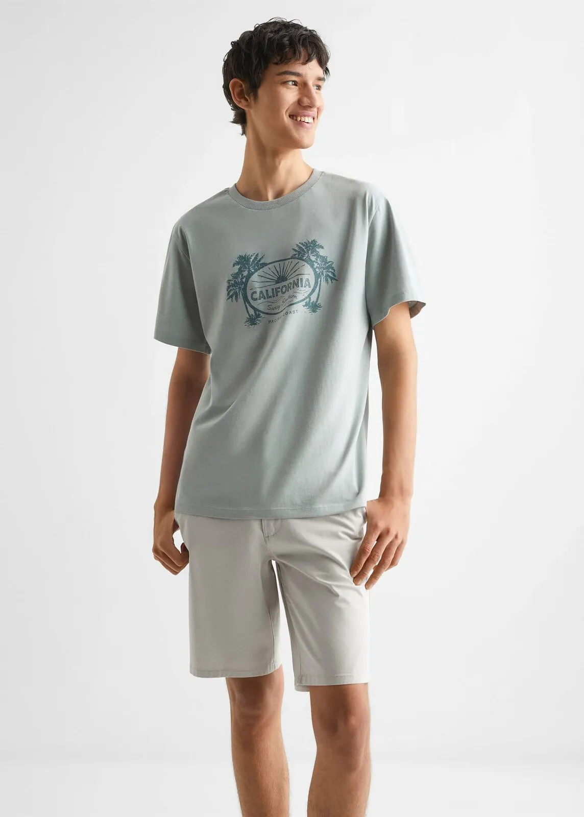 Model in a sage green T-shirt with a “California” palm tree print and light shorts, for Mango