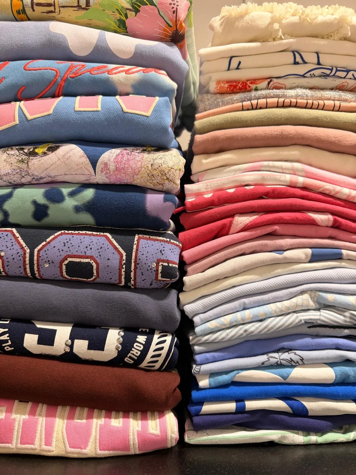 Two stacks of folded sweatshirts and T-shirts with prints, varsity lettering and embroidery